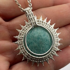 K&ouml;nnte beinhalten: Eine silberne Anh&auml;ngerkette mit einem runden, hellt&uuml;rkisen Stein. Der Stein ist von einem geflochtenen Silberdrahtrahmen mit strahlenf&ouml;rmigen Spitzen umgeben, der einer Sonne &auml;hnelt. Die Kette ist ebenfalls silberfarben.