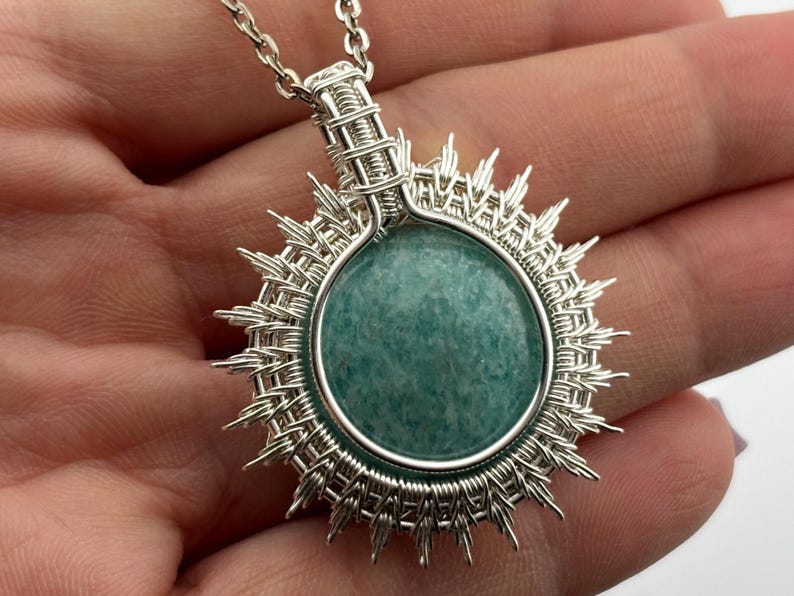 K&ouml;nnte beinhalten: Eine silberne Anh&auml;ngerkette mit einem runden, hellt&uuml;rkisen Stein. Der Stein ist von einem geflochtenen Silberdrahtrahmen mit strahlenf&ouml;rmigen Spitzen umgeben, der einer Sonne &auml;hnelt. Die Kette ist ebenfalls silberfarben.