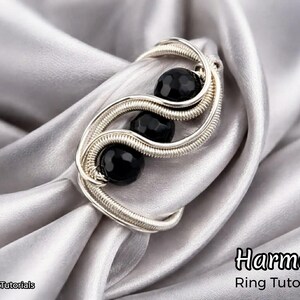 Puede incluir: Un anillo de alambre plateado con tres cuentas negras, sobre un fondo de tela gris plateado drapeada. El diseño del anillo presenta una forma fluida y orgánica. La palabra "Harmony" es visible.