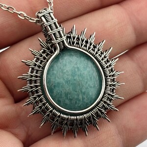 K&ouml;nnte beinhalten: Halskette mit handgefertigtem Silberanh&auml;nger und einem runden, hellblauen Edelstein. Der Edelstein ist von einem Sonnenstrahl-Design aus geflochtenem Silberdraht umgeben. Der Anh&auml;nger h&auml;ngt an einer filigranen Silberkette, ein einzigartiges Schmuckst&uuml;ck.