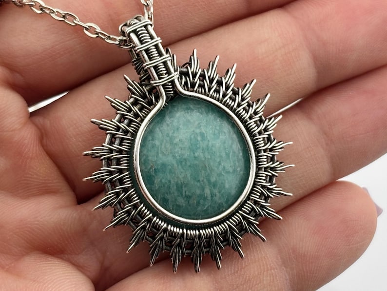 K&ouml;nnte beinhalten: Halskette mit handgefertigtem Silberanh&auml;nger und einem runden, hellblauen Edelstein. Der Edelstein ist von einem Sonnenstrahl-Design aus geflochtenem Silberdraht umgeben. Der Anh&auml;nger h&auml;ngt an einer filigranen Silberkette, ein einzigartiges Schmuckst&uuml;ck.