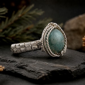 May include: A ring featuring a smooth, round, sage green stone set in a silver wire-wrapped frame. The band is constructed from woven silver wire, creating a textured, basket-weave pattern that encircles the finger, with twisted wire accents.