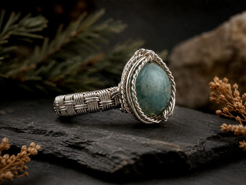 May include: A ring featuring a smooth, round, sage green stone set in a silver wire-wrapped frame. The band is constructed from woven silver wire, creating a textured, basket-weave pattern that encircles the finger, with twisted wire accents.
