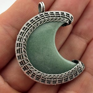 Peut inclure: Un pendentif en forme de croissant avec un centre en pierre vert clair, encadr&eacute; par un fil d'argent tiss&eacute;. Le pendentif a une boucle en haut pour une cha&icirc;ne. Le design est complexe et d&eacute;taill&eacute;, mettant en valeur le contraste.