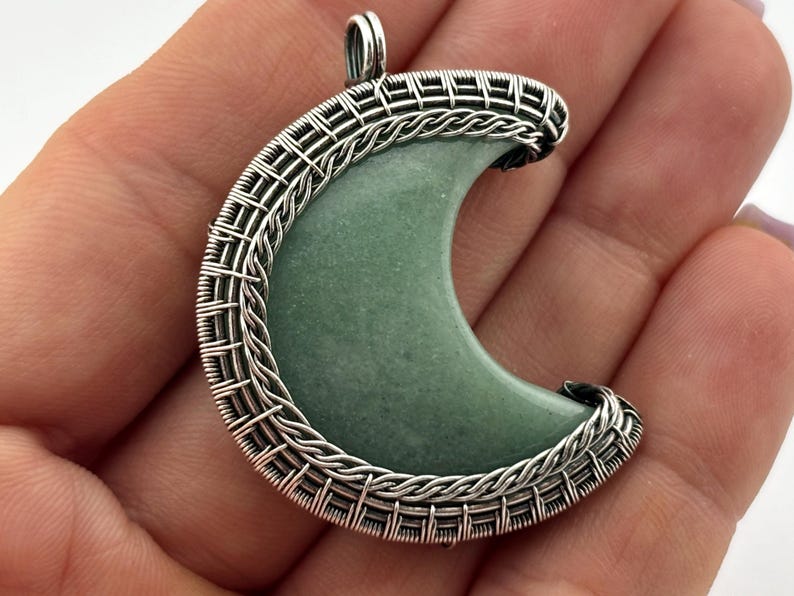 Peut inclure: Un pendentif en forme de croissant avec un centre en pierre vert clair, encadr&eacute; par un fil d'argent tiss&eacute;. Le pendentif a une boucle en haut pour une cha&icirc;ne. Le design est complexe et d&eacute;taill&eacute;, mettant en valeur le contraste.