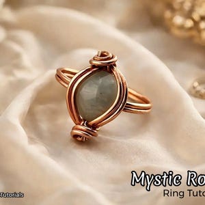 May include: A handcrafted copper wire ring with a pale green oval gemstone. The ring has detailed wire wrapping around the stone and on the band. The text "Mystic Rose Ring Tutorial" is visible.