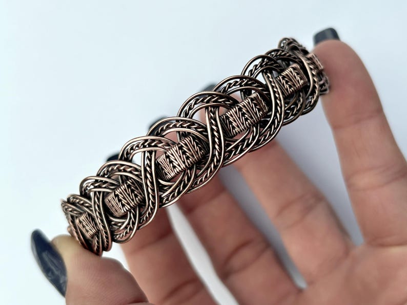 May include: A copper wire bracelet with a woven knot design. The bracelet is made of multiple strands of wire that are intertwined to create a complex pattern.
