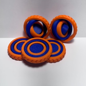 May include: A collection of orange and blue 3D-printed fidget toys. The toys include two sets of interlocking rings and three flat circular discs. The color scheme is a vibrant combination of orange and blue, creating a visually appealing design.