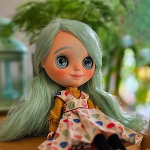 Puede incluir: Una muñeca Blythe con cabello verde menta y ojos grandes y expresivos. La muñeca lleva un top amarillo mostaza debajo de un vestido blanco con estampados coloridos. Calcetines negros y zapatos marrones completan el atuendo. La muñeca está sentada sobre una superficie de madera.