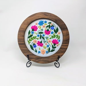 May include: A round embroidery hoop with a white fabric background featuring a colorful floral design. The hoop is made of dark wood and is displayed on a black metal stand.