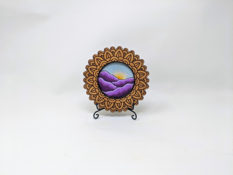 K&ouml;nnte beinhalten: Ein Holzrahmen im Mandala-Stil mit einer violetten und blauen Landschaftsszene im Inneren. Der Rahmen steht auf einem schwarzen St&auml;nder.