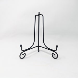 K&ouml;nnte beinhalten: Schwarzer Metall-Pr&auml;sentationsst&auml;nder mit drei Beinen und einem geschwungenen Design. Der St&auml;nder ist ideal zum Pr&auml;sentieren von Tellern, Fotos oder anderen kleinen Gegenst&auml;nden.