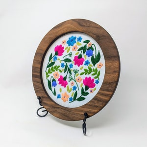 K&ouml;nnte beinhalten: Ein runder Stickrahmen mit wei&szlig;em Stoffhintergrund, der mit einem farbenfrohen Blumendesign verziert ist. Der Rahmen ist aus dunklem Holz gefertigt und auf einem schwarzen Metallst&auml;nder ausgestellt.