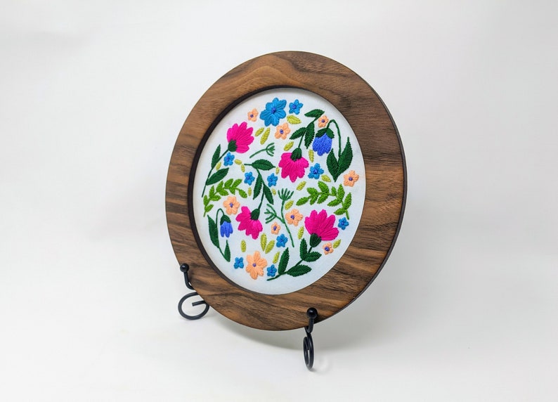 K&ouml;nnte beinhalten: Ein runder Stickrahmen mit wei&szlig;em Stoffhintergrund, der mit einem farbenfrohen Blumendesign verziert ist. Der Rahmen ist aus dunklem Holz gefertigt und auf einem schwarzen Metallst&auml;nder ausgestellt.