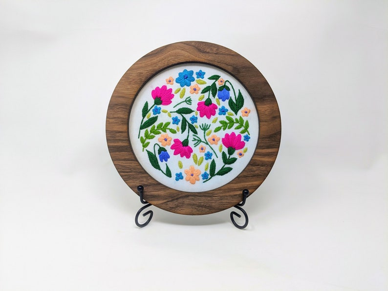 K&ouml;nnte beinhalten: Ein runder Stickrahmen mit wei&szlig;em Stoffhintergrund, der ein farbenfrohes Blumenmuster zeigt. Der Rahmen ist aus dunklem Holz und wird auf einem schwarzen Metallst&auml;nder pr&auml;sentiert.