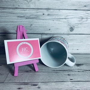 May include: A white ceramic mug with a pink and white design that says "It's more than a creation, it's a dream come true". The mug is sitting on a wooden surface next to a pink easel with a pink business card that says "HC Her Creations".