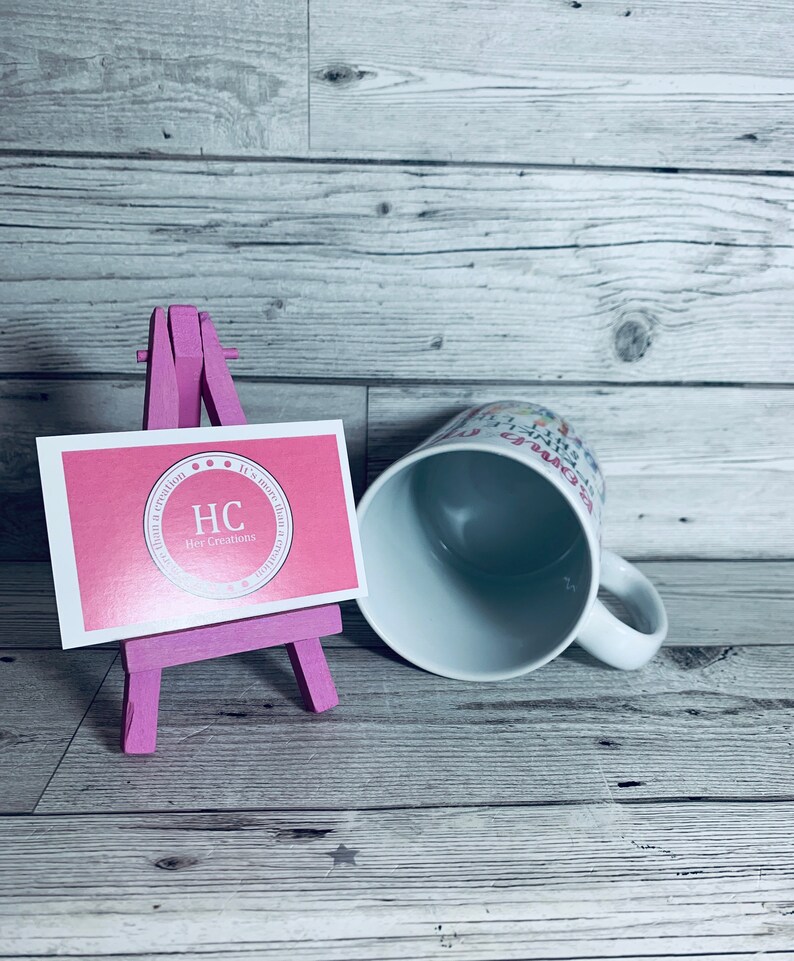 May include: A white ceramic mug with a pink and white design that says "It's more than a creation, it's a dream come true". The mug is sitting on a wooden surface next to a pink easel with a pink business card that says "HC Her Creations".