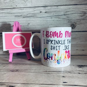 May include: White ceramic mug with a pink and black design. The mug says "I bomb my shit like confetti".