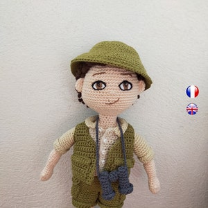 May include: A crocheted doll wearing a green hat, vest, and shorts. The doll has brown hair, brown eyes, and a smile. The doll is holding a pair of binoculars.