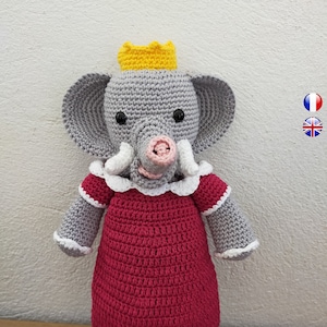 Könnte beinhalten: Ein grauer gehäkelter Elefant, der ein rotes Kleid mit weißen Verzierungen, eine gelbe Krone und rosa Schuhe trägt.