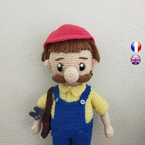 Puede incluir: Una muñeca de crochet con un sombrero rojo, un mono azul y una camisa amarilla. La muñeca tiene el pelo marrón, un bigote marrón y un cinturón de herramientas marrón.