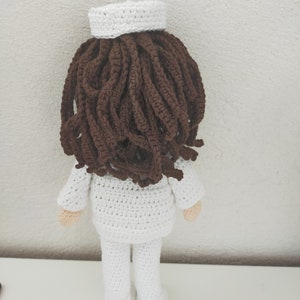 May include: A crocheted doll wearing a white uniform and a white hat. The doll has brown hair and is standing on a white surface.