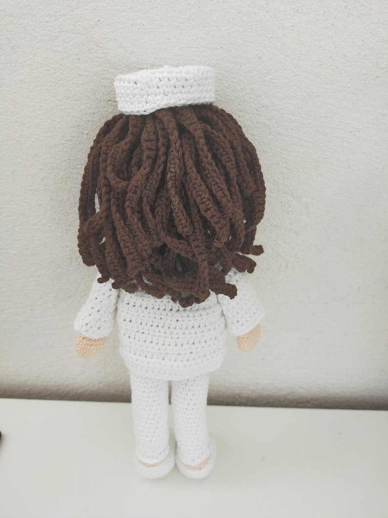 May include: A crocheted doll wearing a white uniform and a white hat. The doll has brown hair and is standing on a white surface.