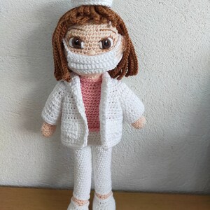 May include: A crocheted doll wearing a white doctor's coat, pants, and a white cap. The doll has brown hair, a pink shirt, and a white face mask.