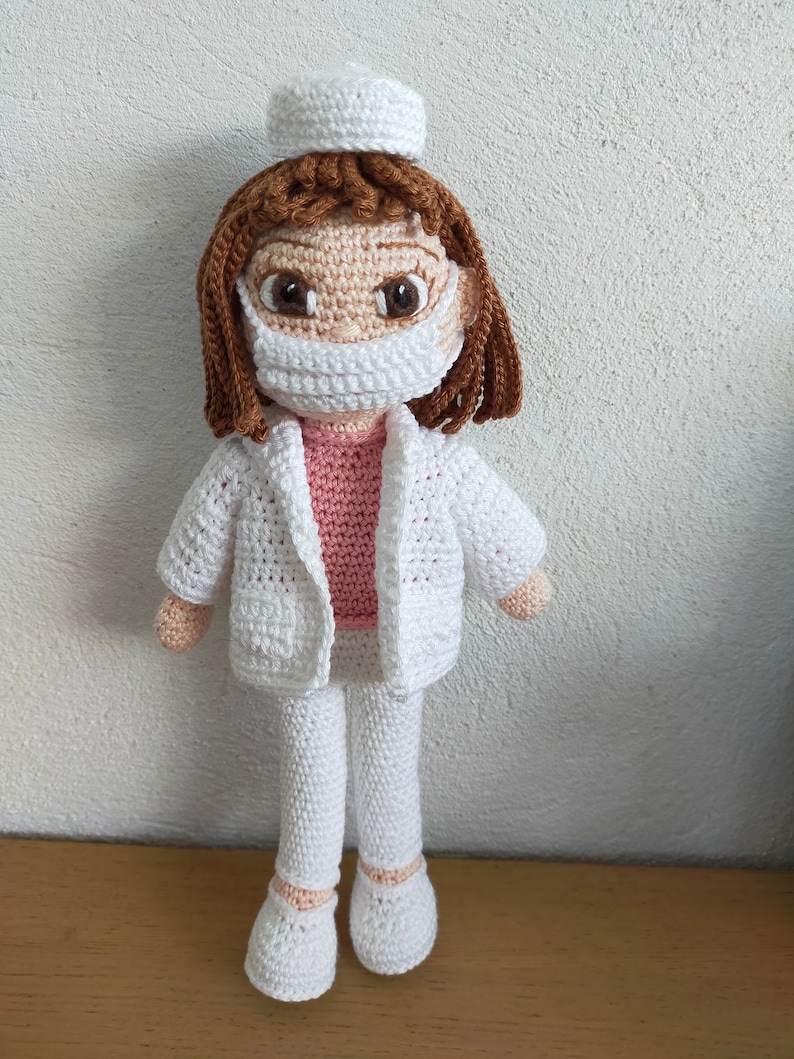 May include: A crocheted doll wearing a white doctor's coat, pants, and a white cap. The doll has brown hair, a pink shirt, and a white face mask.