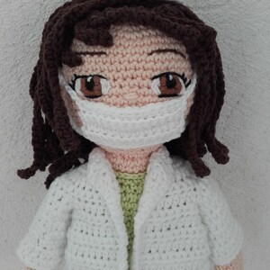 May include: A crocheted doll wearing a white doctor's coat, a white face mask, and a white hat. The doll has brown hair and brown eyes.
