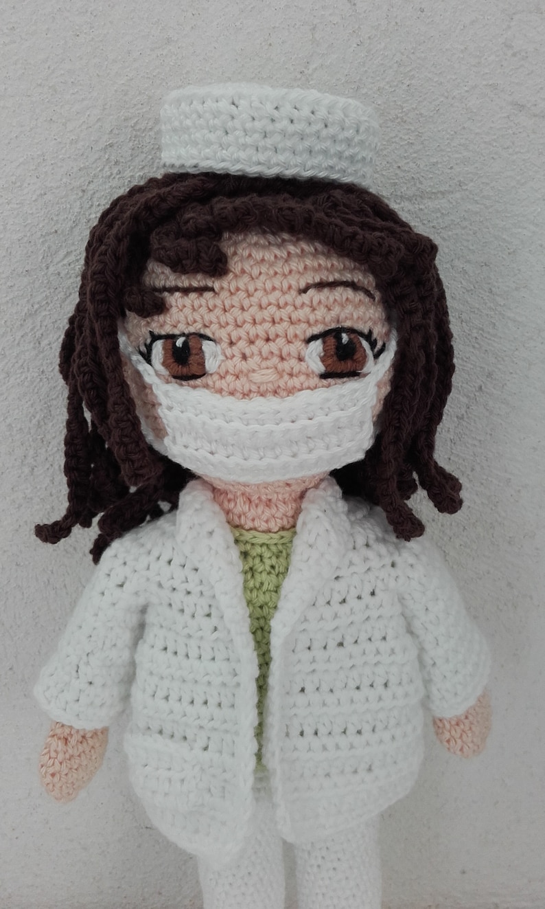 May include: A crocheted doll wearing a white doctor's coat, a white face mask, and a white hat. The doll has brown hair and brown eyes.