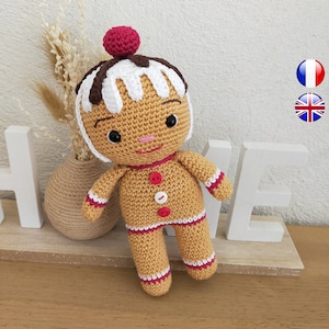 May include: A crocheted gingerbread person doll with a white and brown icing hat and a red cherry on top. The doll has brown eyes, a pink nose, and a smiling mouth. The doll is wearing a brown crocheted outfit with white and red stripes.