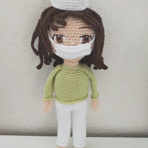 May include: A crocheted doll wearing a white hat, green shirt, white pants, and a white face mask. The doll has brown hair and brown eyes.