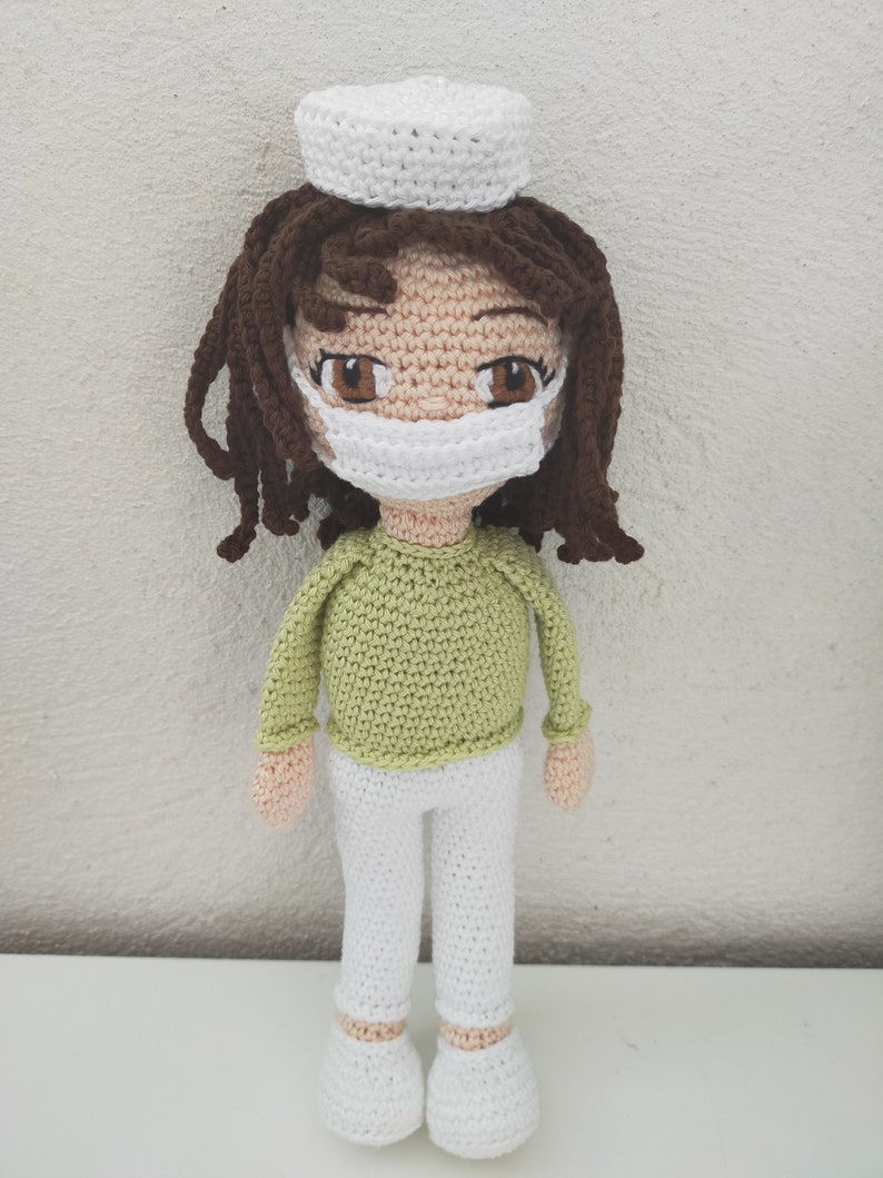 May include: A crocheted doll wearing a white hat, green shirt, white pants, and a white face mask. The doll has brown hair and brown eyes.
