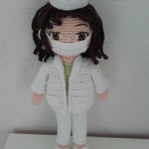 May include: A crocheted doll wearing a white doctor's coat, pants, and a white hat. The doll has brown hair, a light pink face, and a white face mask.