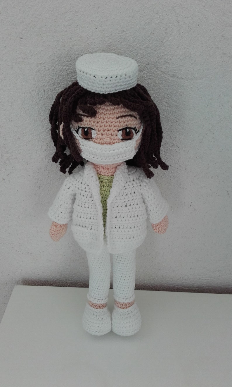 May include: A crocheted doll wearing a white doctor's coat, pants, and a white hat. The doll has brown hair, a light pink face, and a white face mask.