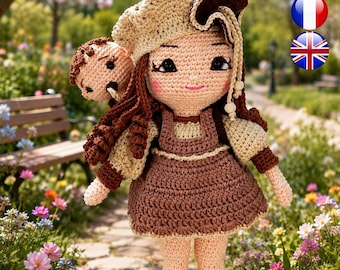 Puede incluir: Una muñeca de ganchillo con largo cabello castaño, que lleva una boina beige, un vestido de peto marrón sobre una blusa beige y zapatos beige. La muñeca tiene una pequeña cabeza de ganchillo redonda y clara unida a la espalda por un cordón.