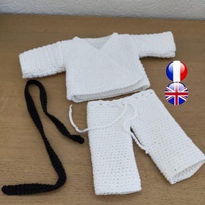 May include: A white crochet baby outfit, comprising a wrap top, trousers, and a black belt. The outfit is handmade with a textured stitch. The top has long sleeves and a V-neck. The trousers have a drawstring waist. The set is displayed on a wooden surface.