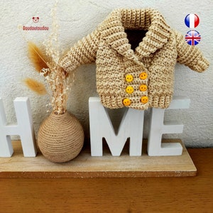 Op de afbeelding: Een gehaakt beige vest met een kraag, gele knopen en een wit houten "HOME" bord.