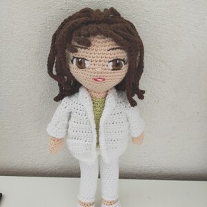 May include: A crocheted doll with brown hair, wearing a white jacket, pants, and shoes. The doll has a green shirt underneath the jacket.