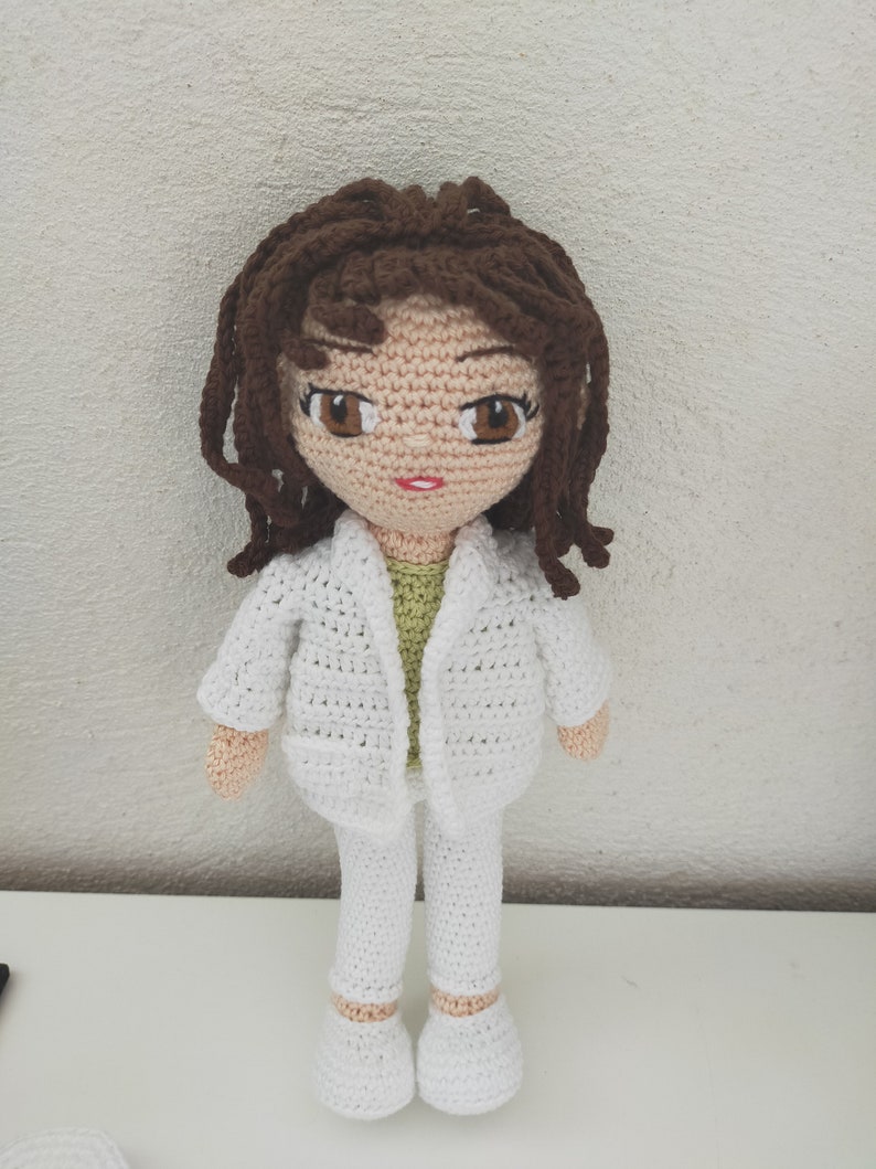 May include: A crocheted doll with brown hair, wearing a white jacket, pants, and shoes. The doll has a green shirt underneath the jacket.