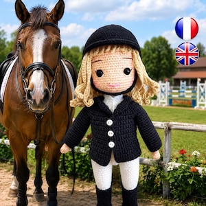 May include: A crochet doll dressed as an equestrian, wearing a black helmet, a black jacket with three white buttons, white breeches, and black boots, standing next to a brown horse with a black bridle and saddle in an outdoor riding arena.