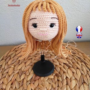 May include: A crocheted doll head with light peach skin, black embroidered eyes, and light brown yarn hair styled with bangs. The head is mounted on a black metal display stand. The text Doudoutoudou is visible in the upper left corner.