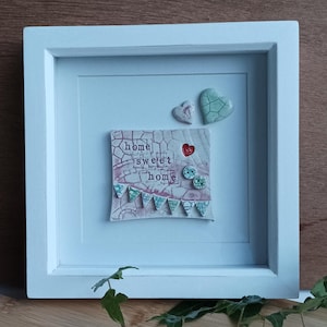May include: A white framed picture with a pink and green ceramic tile that says "home sweet home" with small green and red hearts and flags around it.