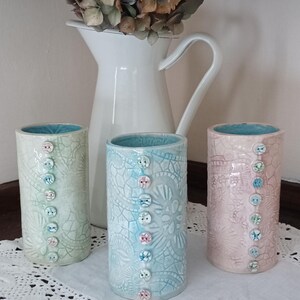 May include: Three ceramic vases with a lace-like design and buttons. The vases are in shades of blue, green, and pink.