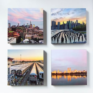 May include: Four square canvas prints featuring New York City scenes. The images include a cityscape at sunset, a view of the skyline from a pier, a train yard with multiple trains, and a reflection of buildings on a lake.