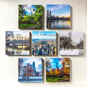 May include: Seven square canvas prints of New York City scenes. Images include Central Park, bridges, cityscapes, and winter scenes. The prints are displayed on a white wall, showcasing the vibrant colors of the city.