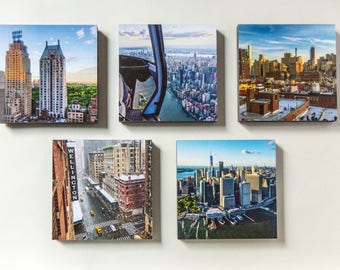 Può includere: Cinque stampe su tela quadrate raffiguranti scene di New York City. Le immagini includono viste aeree di grattacieli, strade cittadine e una vista da un elicottero. Le stampe sono montate su una parete bianca.
