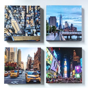 May include: Four square canvas prints of New York City scenes. Images include an aerial view of the Empire State Building, a waterfront view of the city skyline, a street scene with yellow taxis, and Times Square at night.