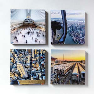 May include: Four square canvas prints featuring New York City scenes. Images include the interior of a modern building, a helicopter view of the city skyline, an aerial view of the Empire State Building, and a train yard at sunset.